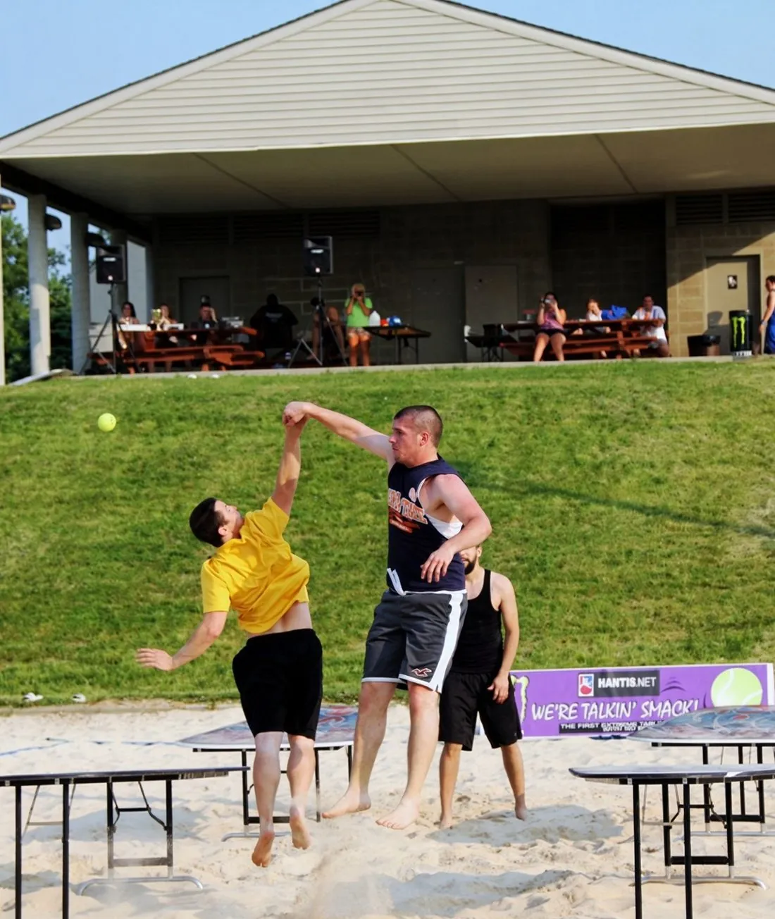 Two players leap for the ball at an outdoor sand-court Hantis tournament as spectators watch from a pavilion
